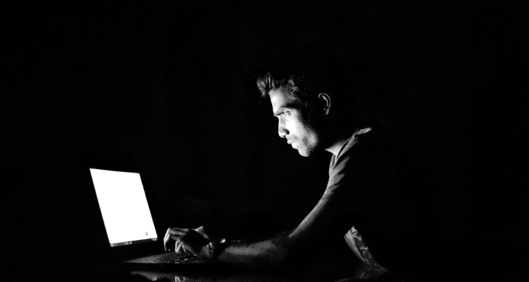 Schwarz-weisses Bild von einem Mann, der in einem dunklen Raum vor einem Laptop. sitzt. Der Bildschirm ist die einzige Lichtquelle