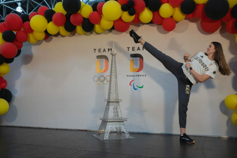 Eine Frau macht in der neuen Team-Kleidung einen High-Kick vor einer dekorierten Wand zu Thema Olympiade 2024