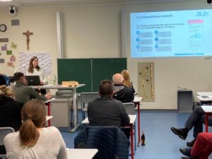 Erwachsene sitzen in einem Klassenzimmer an Einzeltischen. Am Pult steht Anna Leiderer und hält einen Vortrag