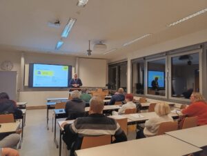 In einem Klassenraum sitzen verteilt Erwachsene Menschen. Am Pult steht Stefan Fäth und hält einen Vortrag